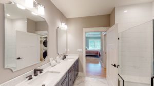 Modern bathroom with dual sinks and large mirror, featuring light fixtures above the sinks. A glass-enclosed shower is on the right, and a door opens to a bedroom in the background.