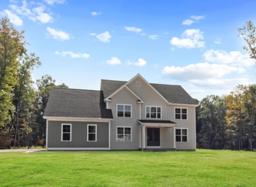 A two-story house with gray siding and a gabled roof stands on a large grassy lawn, surrounded by trees under a partly cloudy blue sky.