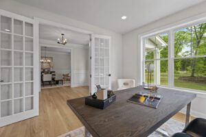 A home office with a dark wood desk, office supplies, and a lamp. French doors open to a dining area with a chandelier. Large windows provide a view of green trees outside.