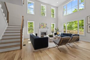 Bright, modern living room with high ceilings, large windows, dark blue sofas, and two accent chairs. A colorful painting hangs above the fireplace, and a staircase is visible on the left.