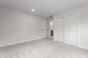 A clean, empty room with beige carpet, white walls, a door to a hallway, and a double-door closet. The ceiling has built-in lighting. A wooden cabinet is visible through the open door.