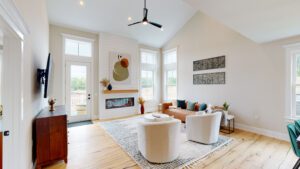 A modern living room with high ceilings features a wall-mounted TV, fireplace, abstract artwork, white armchairs, a brown sofa, and a large window overlooking an outdoor area.