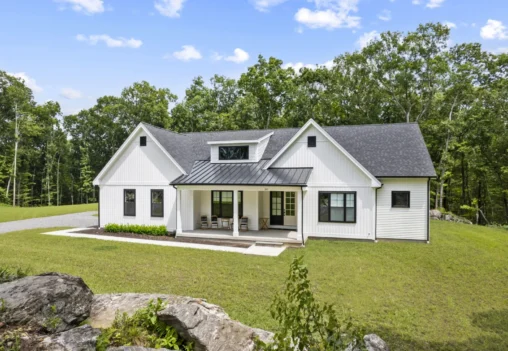 A modern white house with a black roof, nestled in the scenic Willow Creek Estates, surrounded by a lush grassy area with trees and a clear blue sky in the background.