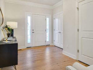 A modern entryway with light hardwood floors, white walls, a white door with windows, a black console table with a lamp, and a small plant. A door to the right leads to another room.