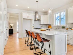 A modern kitchen features a large island with four brown stools, white cabinetry, stainless steel appliances, and pendant lighting. Light wood flooring extends through the room, creating an open, airy feel.