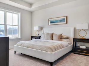 A modern bedroom with a large bed centered between two black nightstands with lamps. A landscape painting hangs above the bed, and a window on the left side allows natural light to brighten the space.