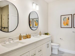 A modern bathroom features a double sink vanity with two circular mirrors, minimalist decor, and framed abstract art on the wall. A toilet is visible in the corner.