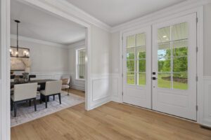 This image shows a foyer with double doors and a view into the adjacent dining room. The foyer features light hardwood flooring and white-paneled walls. The dining room has a dark table and beige chairs.