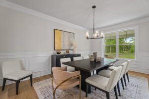 A dining room with a dark rectangular table, beige upholstered chairs, a pink accent chair, modern chandelier, large window, and abstract artwork on the wall.