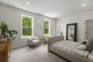 The modern bedroom at 275 Vista View features a large bed, two cozy armchairs, and a stylish floor mirror, all enhanced by lush greenery outside the windows.