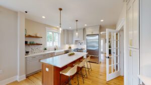 Modern kitchen with stainless steel appliances, an island with seating, pendant lighting, white cabinets, wooden floors, and a French door leading to another room.