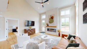 A modern living room with high ceilings, a leather sofa, white armchairs, a round coffee table, a wall-mounted TV, a large fireplace, and contemporary artwork. There is a view into an adjacent bedroom.