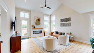 Spacious modern living room with large windows, light-colored walls, wooden flooring, and vaulted ceiling. It includes a fireplace, artwork, a sofa, two armchairs, a rug, and a ceiling fan.