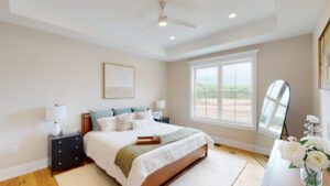 A bedroom with a large window, a bed with white and gray bedding, two nightstands with lamps, and an arched mirror. A ceiling fan is visible above, and a tray with items is on the bed.