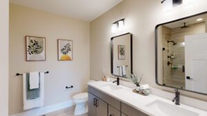 A modern bathroom with double sinks, a large mirror, and light fixtures above. Towels hang on the wall next to framed artwork. A shower with a glass door is visible in the background.