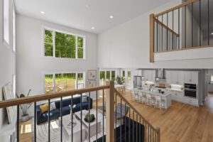 Bright, modern open-concept living area with high ceilings, large windows, a kitchen island, and a loft balcony overlooking a living room furnished with a navy sectional couch.