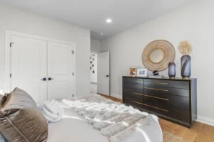 Minimalist bedroom with a cozy bed, dark wood dresser, Summertime vases, and Circle mirror. White walls and doors.