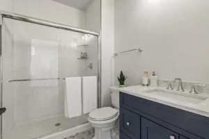 Summertime Circle: Modern bathroom with glass shower, blue vanity, marble countertop, and a small plant on the toilet tank.