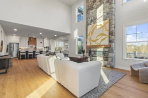 Spacious living room with white sofas, stone fireplace, and modern kitchen; large windows and natural light circling in.