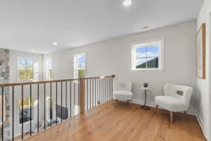 Minimalist loft area with two white chairs, a small table, and wooden floors in Summertime Circle overlooks a stone-accented wall.