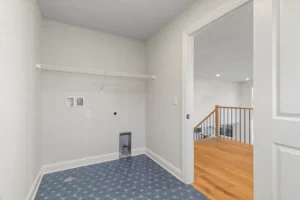 Empty laundry room with blue patterned tile floor, white walls, and wire shelf above utility hookups. Summertime Circle doorway open.