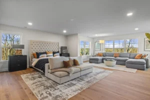 Spacious bedroom with a large bed, beige sofa, and gray sectional couch, featuring wooden floors and large circle windows.