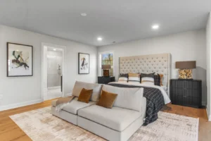 Bedroom with a large bed, Summertime Circle art, beige sofa, and a walk-in shower visible in the background.