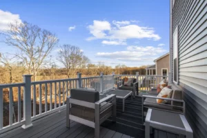 A summertime deck with modern chairs and pillows overlooks a wooded area under a blue sky with clouds.