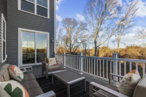 Summertime patio with chairs and table on a wooden deck, offering a view of leafless trees and blue sky beyond the railing.