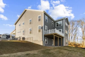 A modern gray house with multiple windows and a balcony on Summertime Circle, set on a grassy slope under a cloudy blue sky.