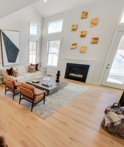 A modern living room features a white sofa, two brown chairs, a wooden coffee table, a fireplace, and abstract wall art. Large windows bring in natural light, and a woven basket sits on the floor.