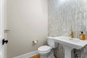 A minimalist bathroom with a white toilet and pedestal sink, gold faucet and soap dispenser, tiled wall, and hardwood floor. A toilet paper holder is mounted on the wall.