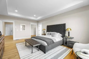 Spacious bedroom with a large dark grey bed, tufted headboard, two nightstands with lamps, a striped rug, and a cushioned bench at the foot of the bed.