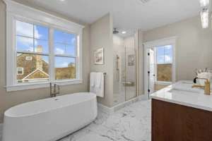 A modern bathroom with a freestanding bathtub, glass shower, double sink vanity, and large window overlooking a house and landscape. The floor is white marble with a gray pattern.