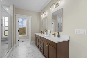 Modern bathroom with double wooden vanity, marble countertop, and dual mirrors. Glass-enclosed shower to the left and large window with a scenic view in the background.