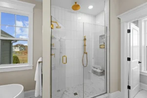 Modern bathroom with a glass shower enclosure, gold fixtures, and white tiles. A window and a towel rack are on the left.