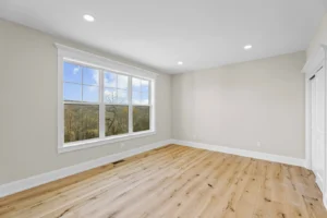 Empty room with wooden floor, beige walls, and a large window displaying an outdoor view.