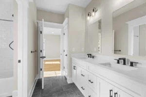 A modern bathroom with dual sinks, a large mirror, black fixtures, white cabinetry, and gray tile flooring. A doorway opens to a sunlit room with hardwood floors.