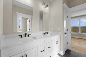 A modern bathroom features a double sink with black fixtures, white countertops, and a large mirror. A window provides natural light, and a view of a room with wood flooring is visible.