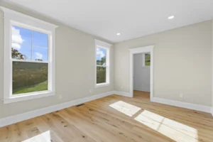 Empty room with light wooden floor, two windows with white trim, beige walls, and a white door, illuminated by natural light.