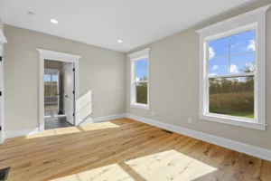 A bright, empty room with light wood flooring, two large windows, and a white door leading to another space. The walls are painted beige.