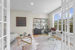 Bright home office with glass desk, modern chair, shelves, and geometric decor; features large window and abstract art on beige walls.