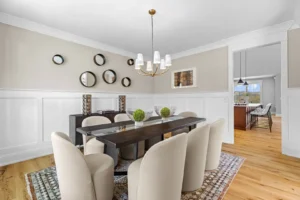 Modern dining room with a dark wooden table, eight beige chairs, and a chandelier. Circular mirrors and decorative items on the wall. Light wood flooring and adjacent view into another room.