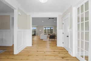 A bright, open-concept living space with light wood floors, white walls, and large windows. A seating area is visible in the background.