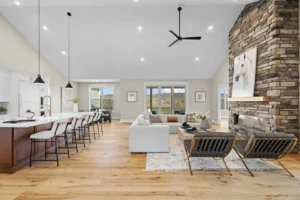 Open-concept living area with a kitchen and island on the left, a white sofa and chairs, and a stone fireplace on the right. Hanging lights and a ceiling fan are above. Large windows in the background.