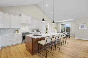 Modern kitchen with a high ceiling, featuring a large island with bar stools, white cabinets, pendant lights, and hardwood floors. A window provides natural light in the background.