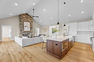 Open-concept living room and kitchen with a stone fireplace, wooden floors, white cabinetry, and a large island. Cozy seating area with a wall decoration above the fireplace.