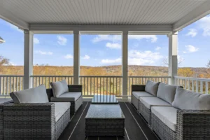 Outdoor seating area with two wicker sofas, chairs, and a glass coffee table on a covered porch. Overlooks a scenic view of autumn trees and hills under a partly cloudy sky.