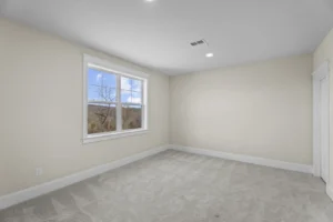 Empty room with beige walls, carpeted floor, a window showing an outdoor view, and a closed door on the right side.