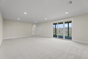 Spacious empty room with beige carpet, sliding glass doors leading to a patio, and recessed ceiling lights. White walls and closed doors are visible.